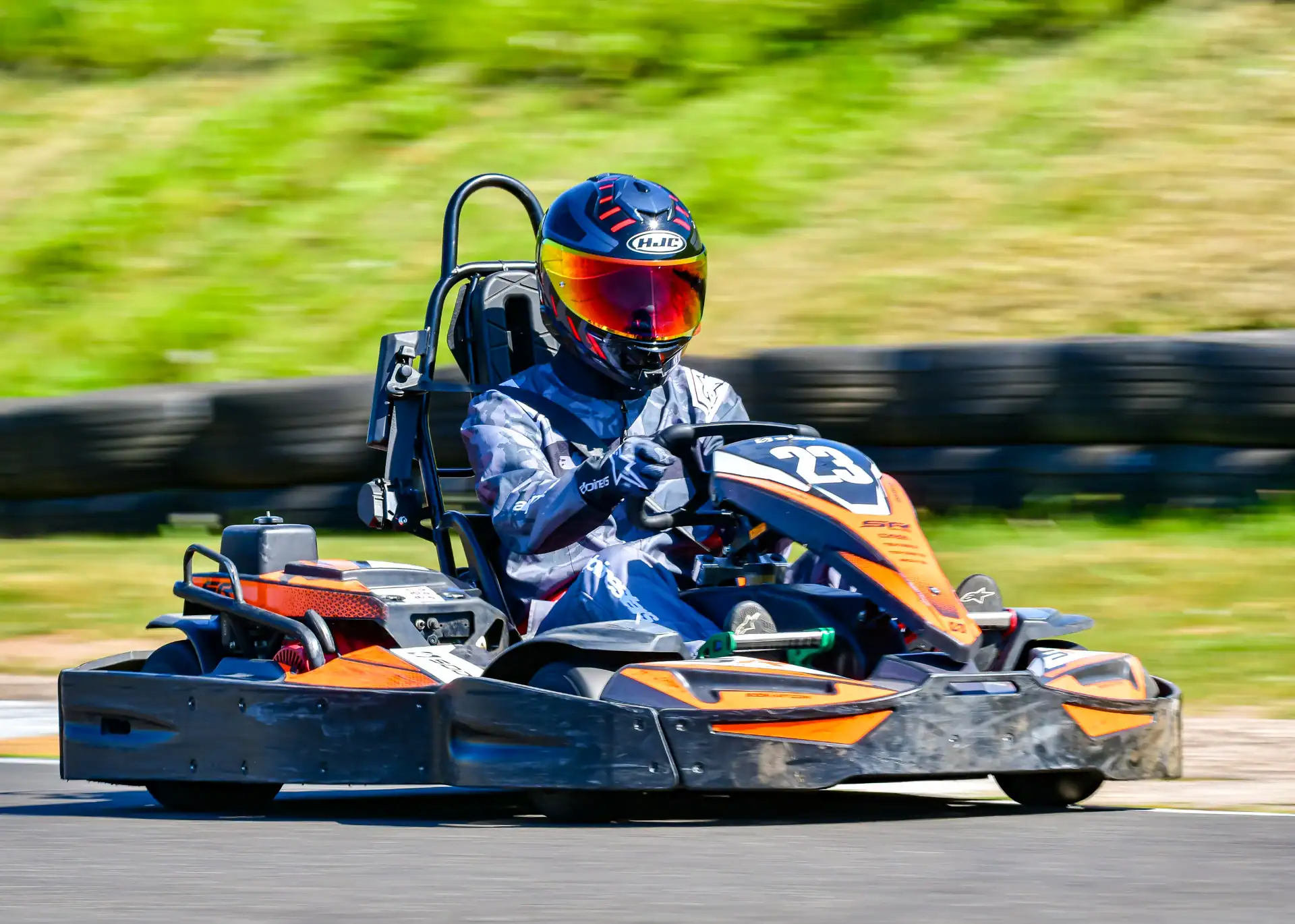 Person wearing a helmet and racing suit drives a go-kart with the number 23 on an outdoor track with a grassy backdrop.