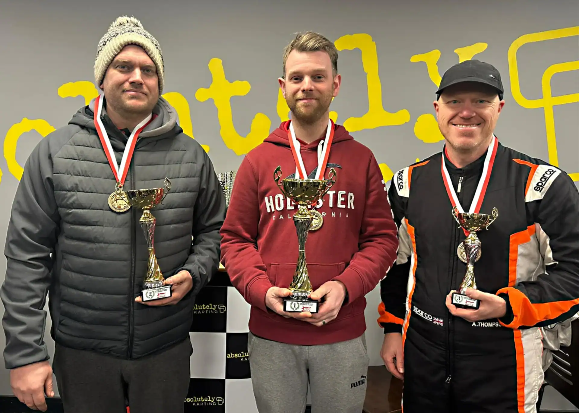 Three men stand indoors holding trophies and wearing medals, posing in front of a chequered backdrop with yellow text.