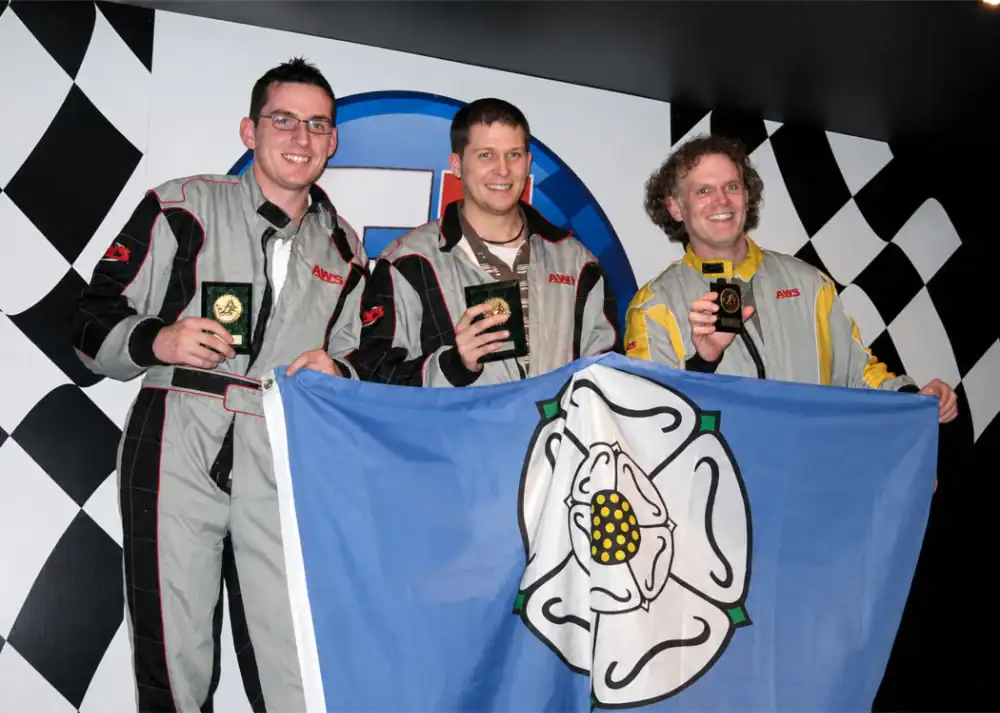Three men in racing suits hold medals and a blue flag with a white rose emblem, standing in front of a black-and-white chequered backdrop.