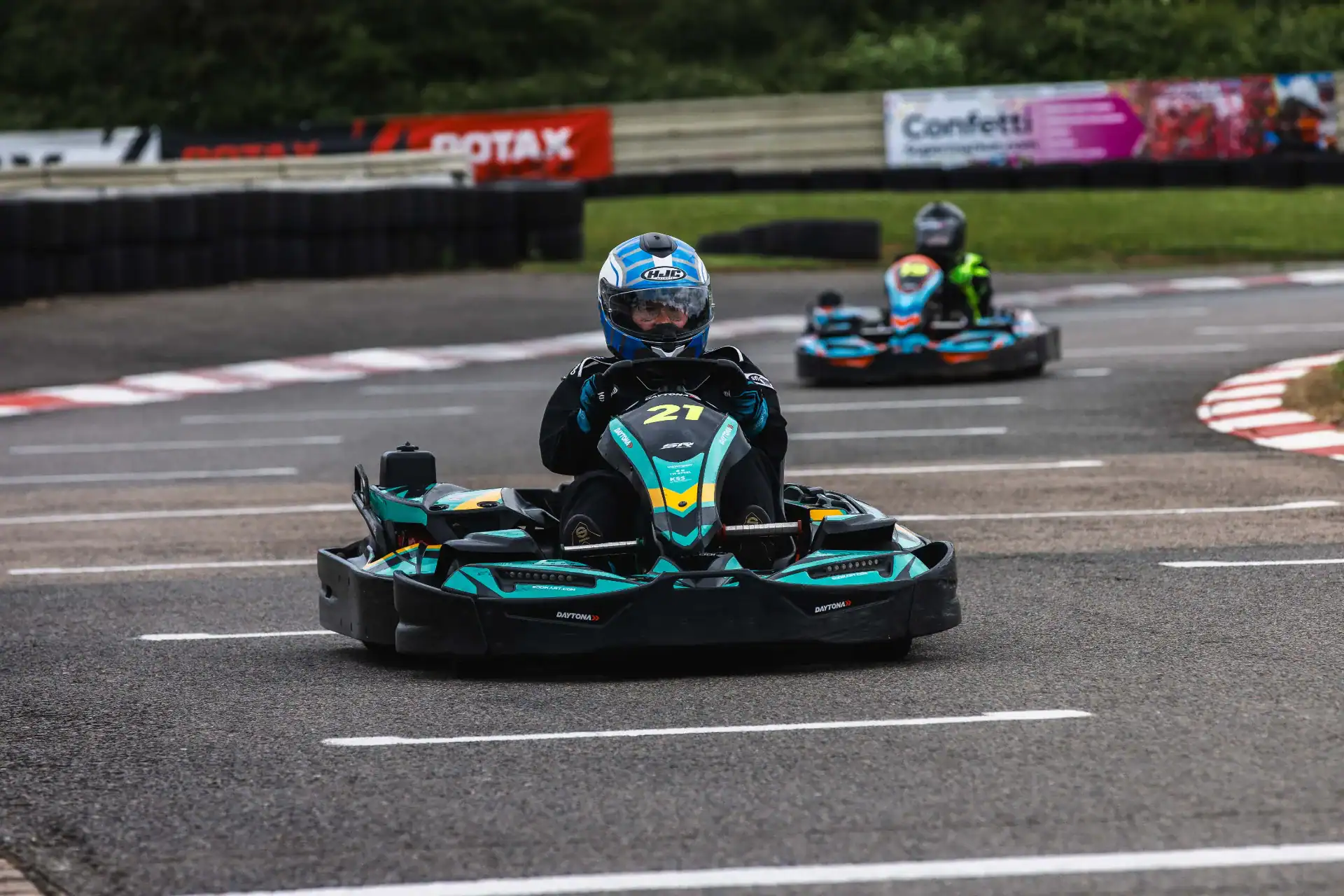 Two people wearing helmets drive go-karts on an outdoor track, with the person in kart number 21 leading.